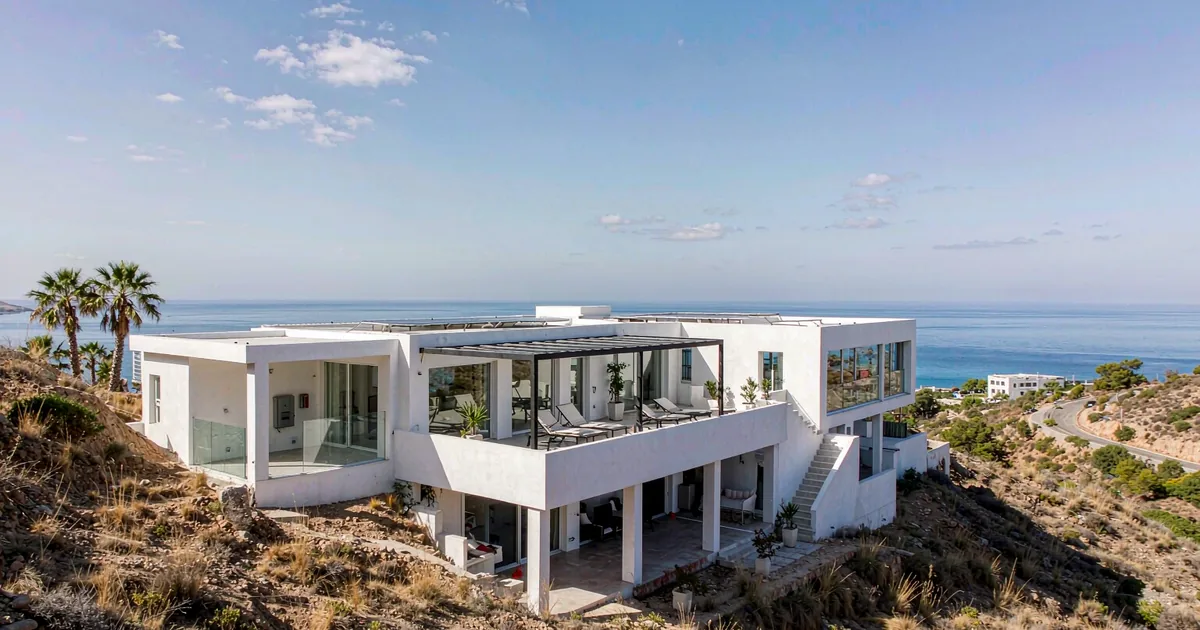 Modern white Mediterranean coastal home with a covered wall-mounted home battery cabinet and rooftop solar, sea in the background
